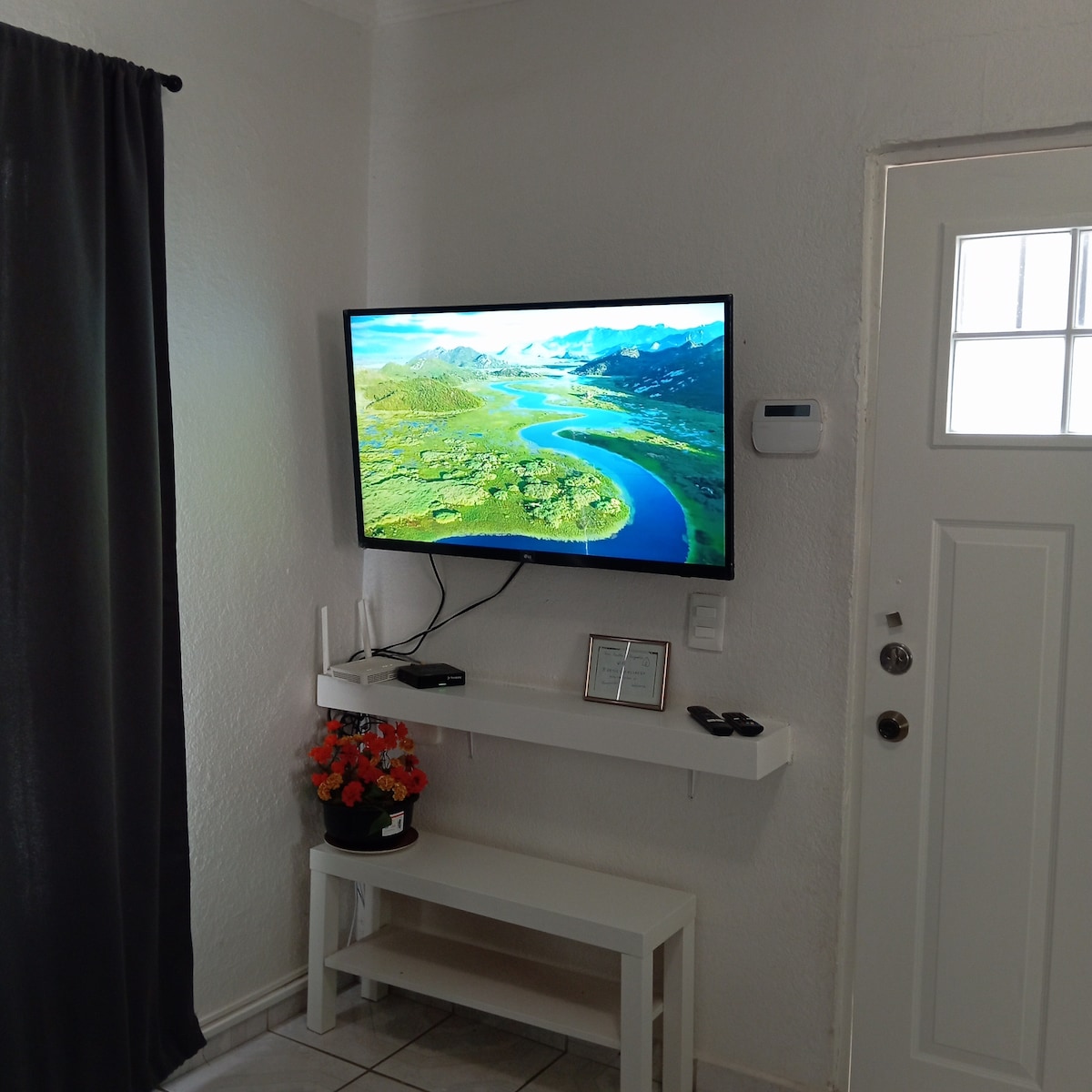 A wall-mounted television displays a vibrant landscape of mountains and rivers. Beneath it, a white shelf holds a small flower pot and remotes. A door with glass panels is visible to the right, complemented by a dark curtain on the opposite wall.