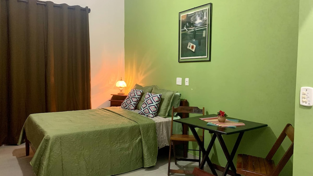 A room features a spacious bed covered with a green bedspread, complemented by patterned pillows. A small table and two wooden chairs are present beside the bed. A warm light emanates from a bedside lamp, and a framed photograph adds a decorative touch to the vibrant green wall.