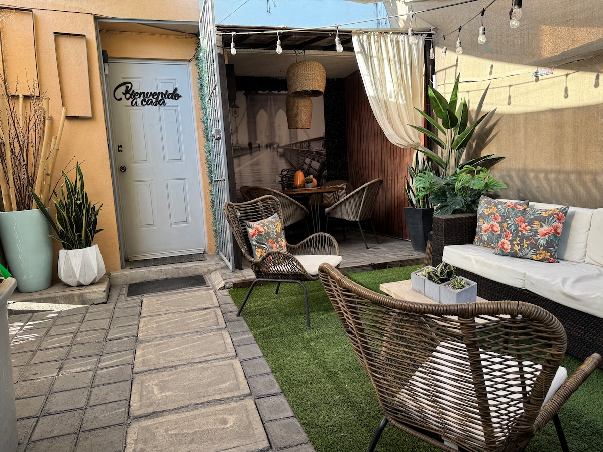 An outdoor seating area features a combination of wicker chairs and a white sofa, arranged on artificial grass. Potted plants add greenery, while string lights hang above for illumination. A door leads into the space, and the wall displays a monochrome photograph of a bridge.