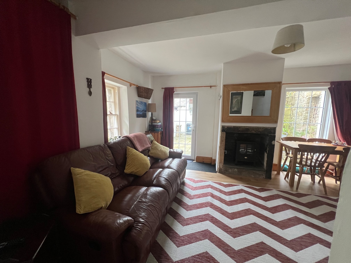 A well-lit living/dining area is showcased, featuring a cozy reclining sofa with yellow cushions and a dining table for four. A fireplace adds charm, while large windows allow natural light to fill the space. A patterned rug complements the room's decor.