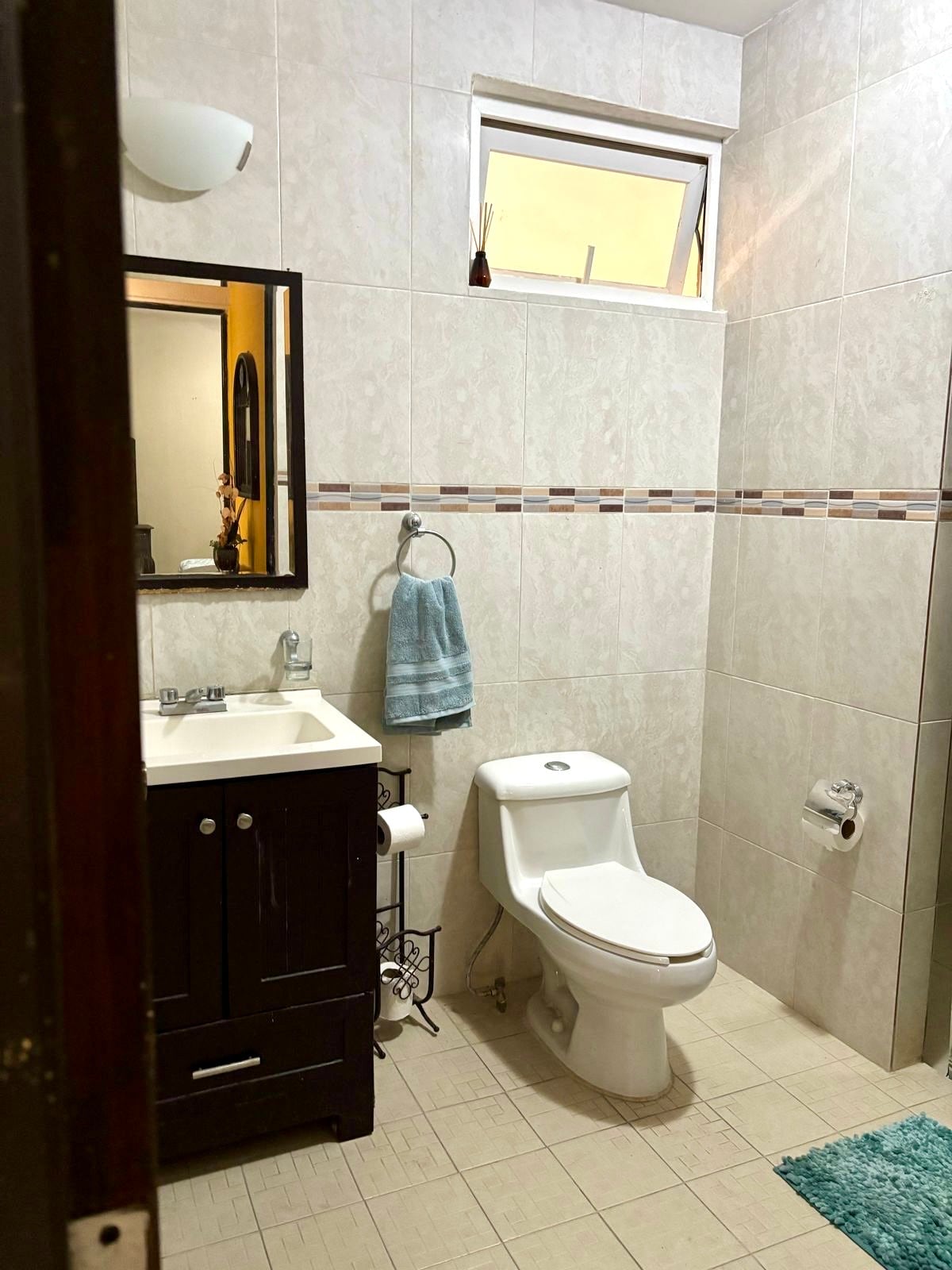 A well-appointed bathroom features light-colored tiles and a combination of dark cabinetry and a white sink. A mirror is mounted above the sink, and a towel rack holds a soft blue towel. Natural light enters through a small, frosted window.