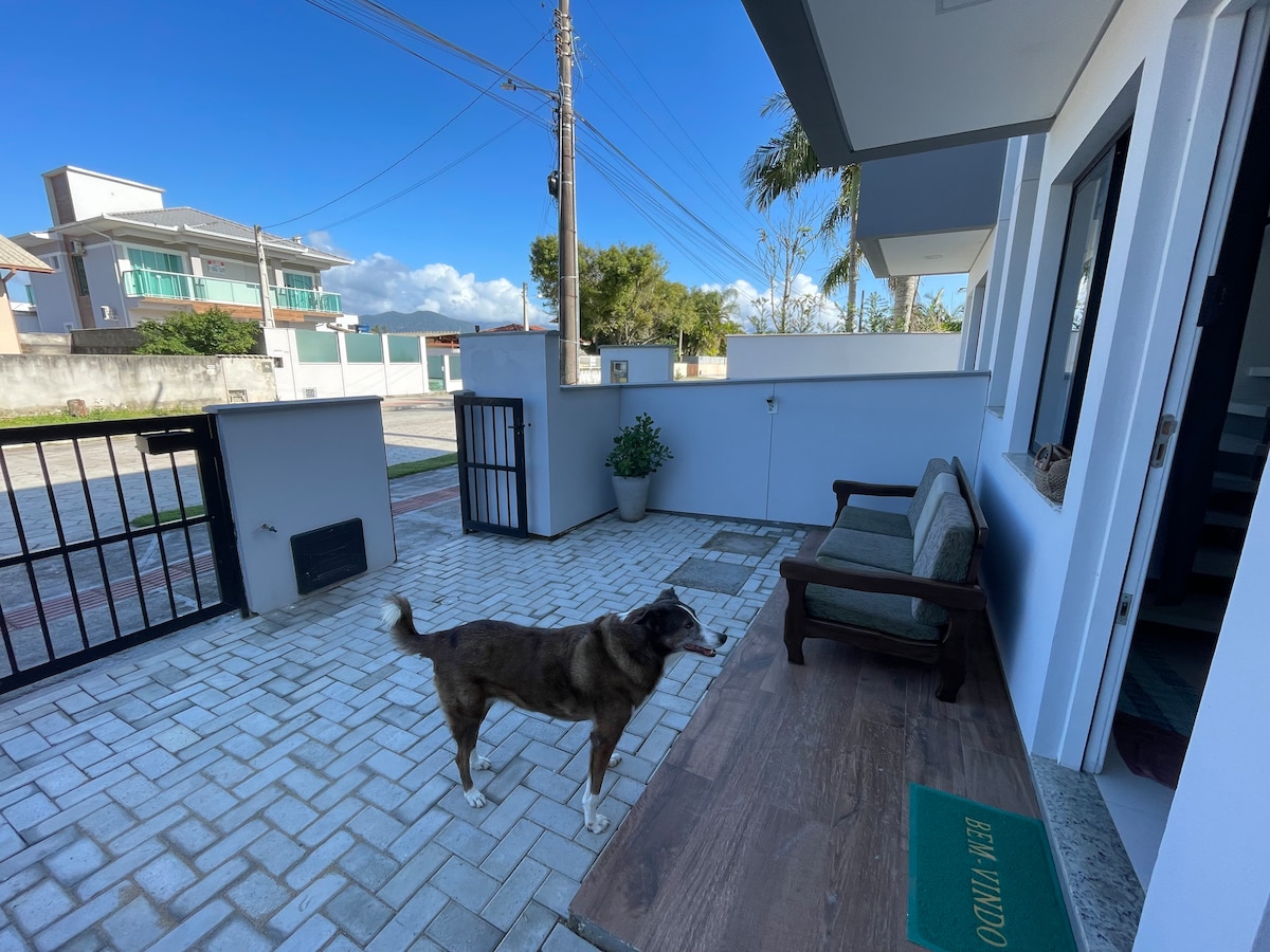 A welcoming entrance features a paved patio with a wooden bench and plant. A pet-friendly dog is seen outdoors, and a secure gate is visible, contributing to privacy. Clear skies enhance the overall setting, suggesting a tranquil environment near the beachfront.