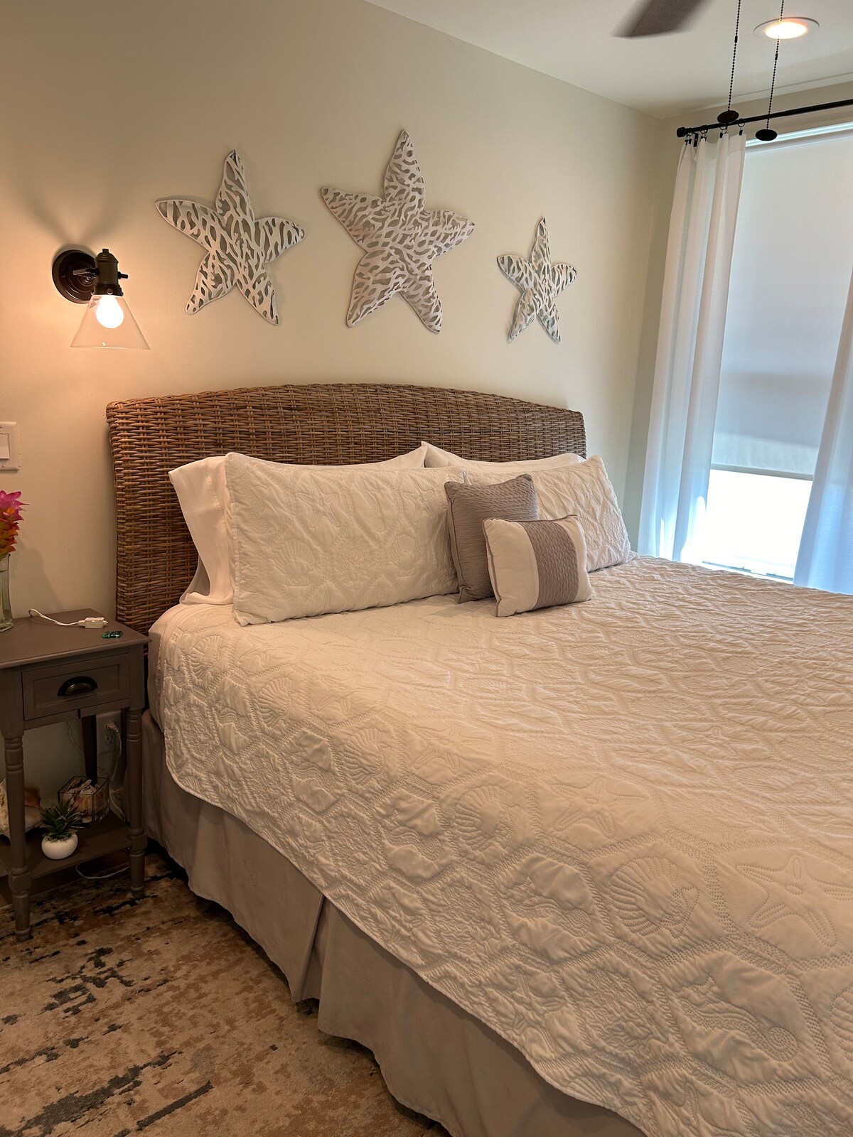 A serene bedroom features a spacious bed with a textured white coverlet, complemented by decorative star motifs on the wall. A small bedside table holds a floral arrangement, while natural light filters through sheer curtains, contributing to a calming ambiance.