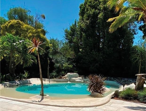 A serene beach pool is framed by lush greenery, including tall palm trees and various shrubs. The pool features a naturalistic design with curved edges, and the clear water reflects the bright blue sky above.