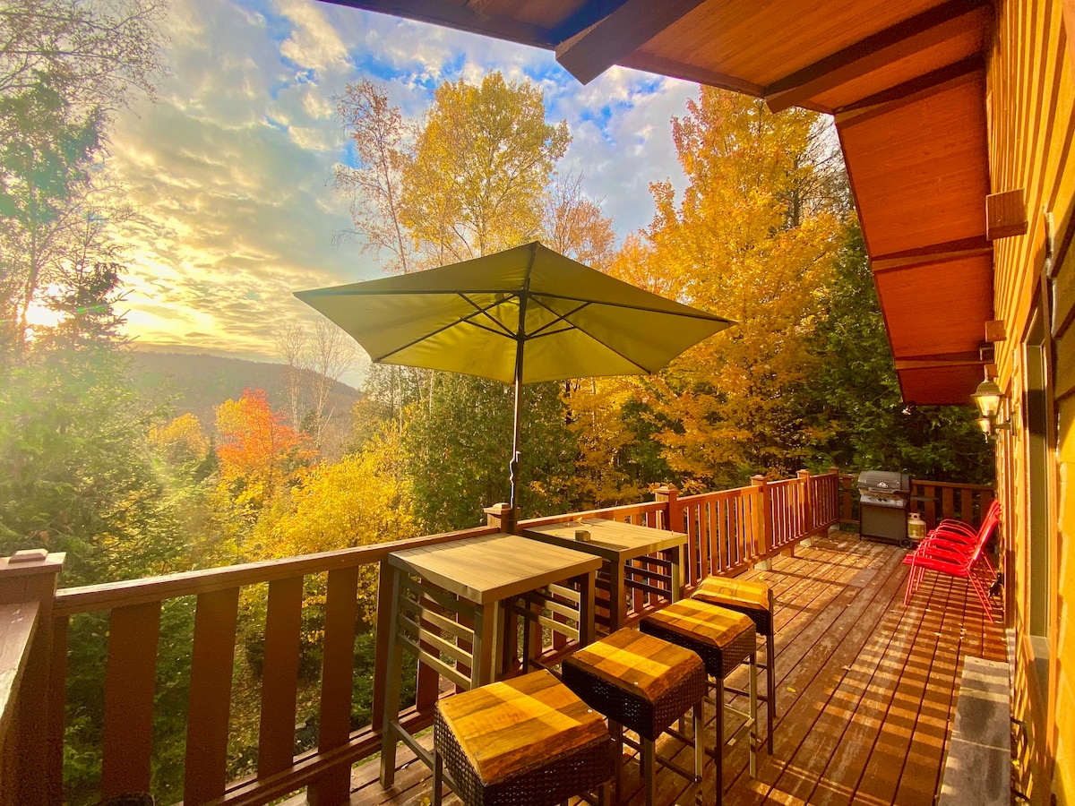 An outdoor deck is shown with a large green umbrella providing shade. Several high-top chairs surround a table, with a gas grill visible nearby. The vibrant autumn foliage adds color to the view, complemented by soft evening light filtering through the trees.