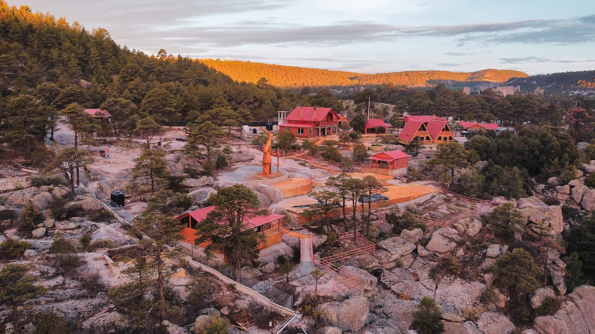 Aerial view of a scenic retreat nestled among rocky terrain and pine trees. Distinctive red-roofed cabins are positioned amidst the natural landscape, surrounded by serene greenery and expansive open space.