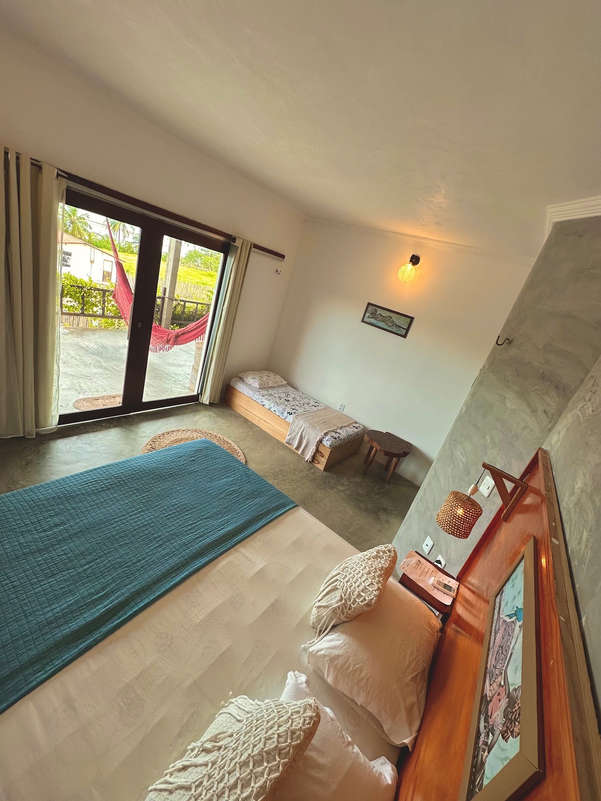 A spacious room is visible, featuring a large bed with decorative pillows. A cozy single bed is positioned beside it. Natural light enters through glass doors leading to a veranda, where a hammock is displayed, creating a relaxed atmosphere.