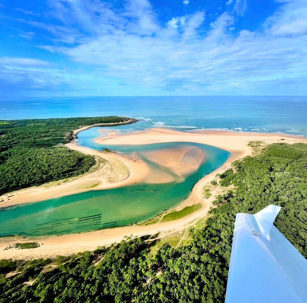 Aerial view of a coastal landscape showcases a winding river flowing into the ocean, with sandy shores and lush greenery. The sky is filled with fluffy clouds, casting shadows over the tranquil waters, creating a serene and captivating vista.