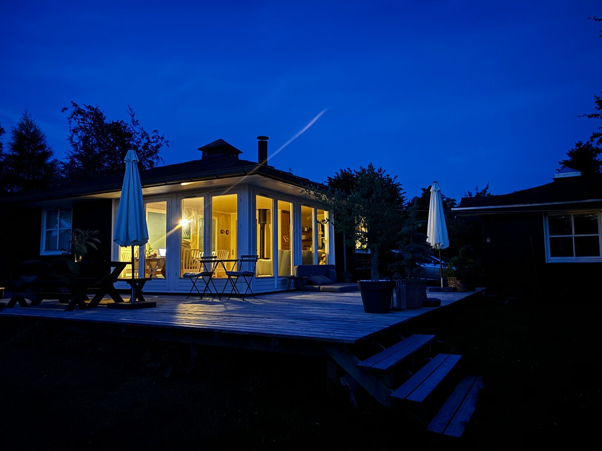 The exterior of the summer house is illuminated at dusk, highlighting a spacious wooden deck. Multiple seating areas are visible, surrounded by plants. Soft light glows from within the house, creating a welcoming and tranquil atmosphere.