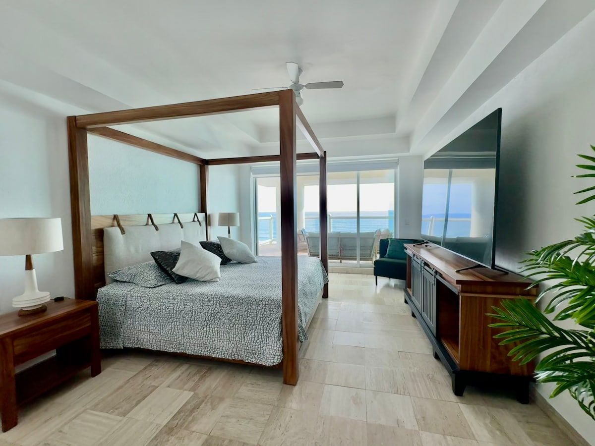 A spacious bedroom is highlighted by a four-poster bed adorned with decorative pillows. A large television is mounted on the wall opposite the bed. Floor-to-ceiling windows offer unobstructed views of the ocean. A cozy seating area is positioned nearby, complemented by natural light.