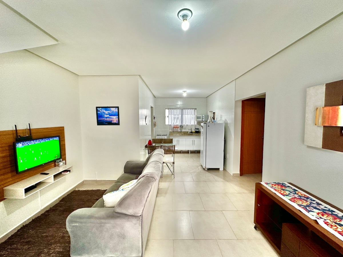 The living area features a comfortable sofa and a wall-mounted HDTV. A minimalistic wooden shelf displays the television, while a bright window allows natural light to fill the space. The kitchen is visible in the background, equipped with essential appliances and a dining space.