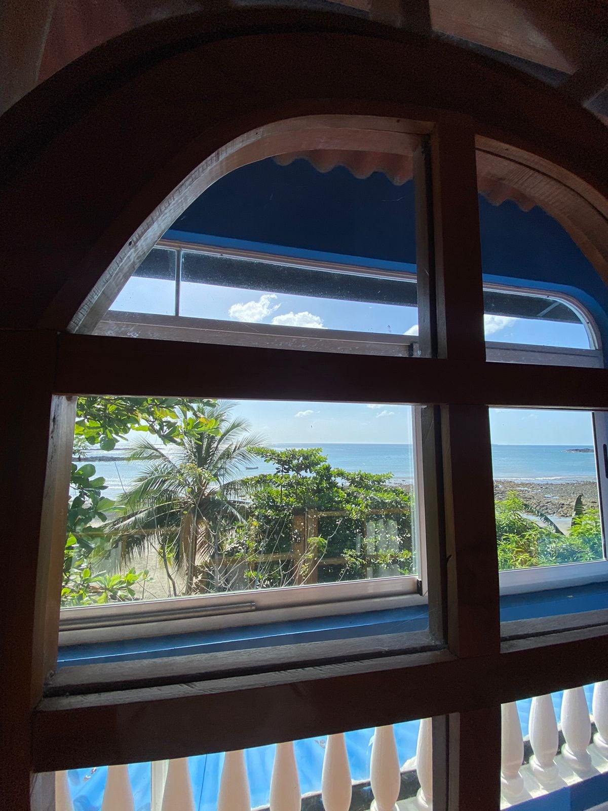 A window framed by curved wooden casements offers a view of the ocean and lush greenery. Palm trees are visible, swaying gently in the breeze. Natural light floods the interior, illuminating the space with a serene ambiance.