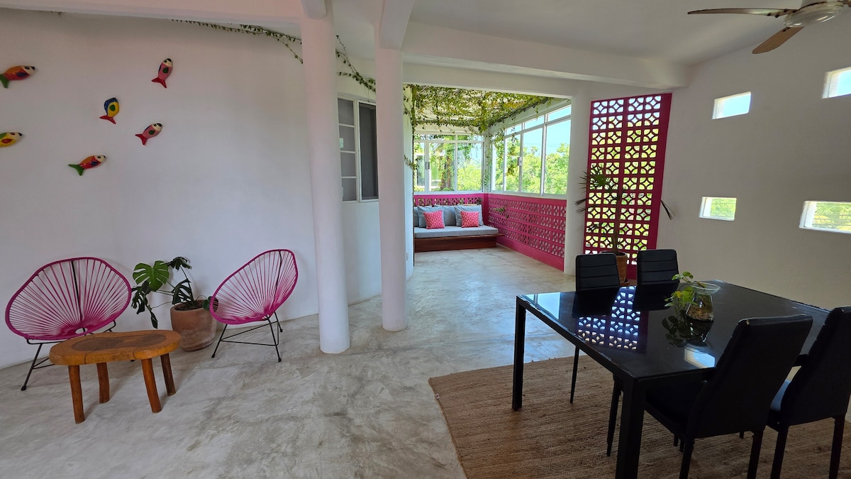 A spacious living area features a dining table for four, complemented by two pink chairs. Natural light fills the space through varied windows, and a cozy corner with cushions invites relaxation. Decorative fish provide a splash of color against the white walls.