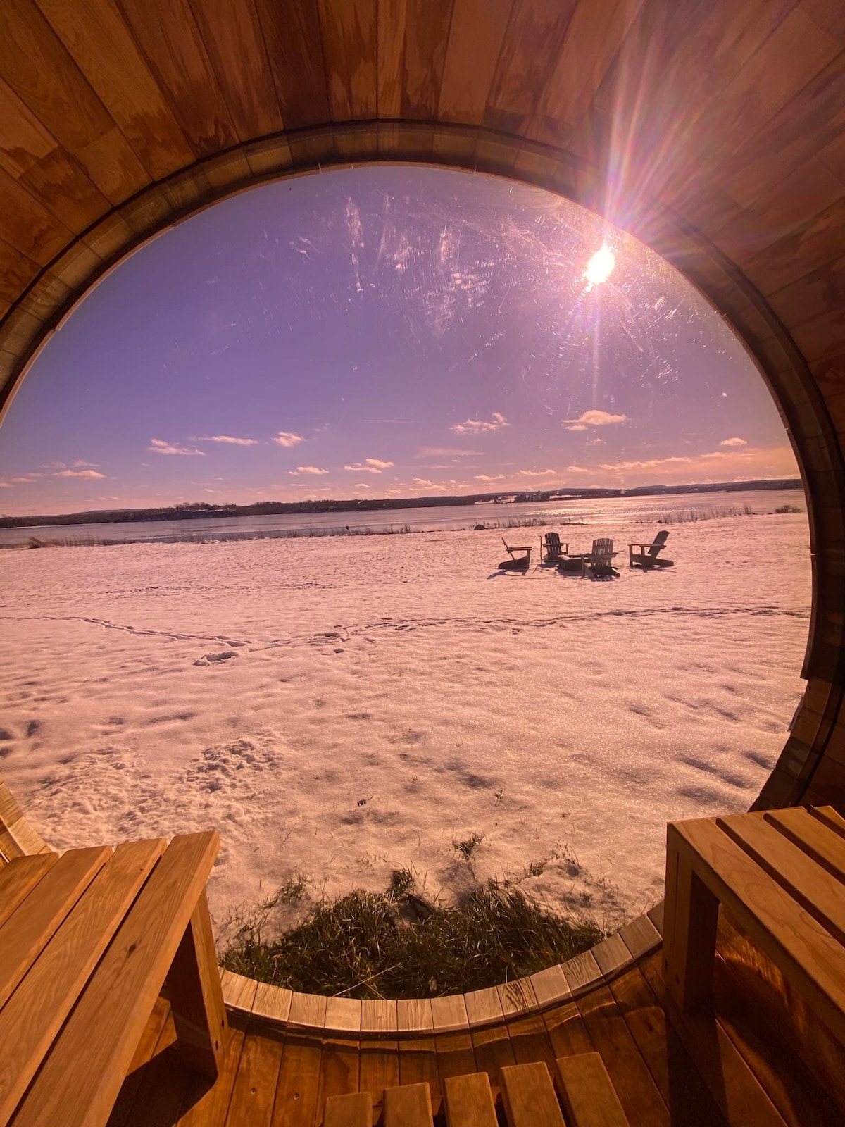 A circular window frames a snowy landscape, revealing a serene view of two Adirondack chairs set on a white blanket of snow. Soft sunlight bathes the scene, highlighting the natural beauty of the surroundings.