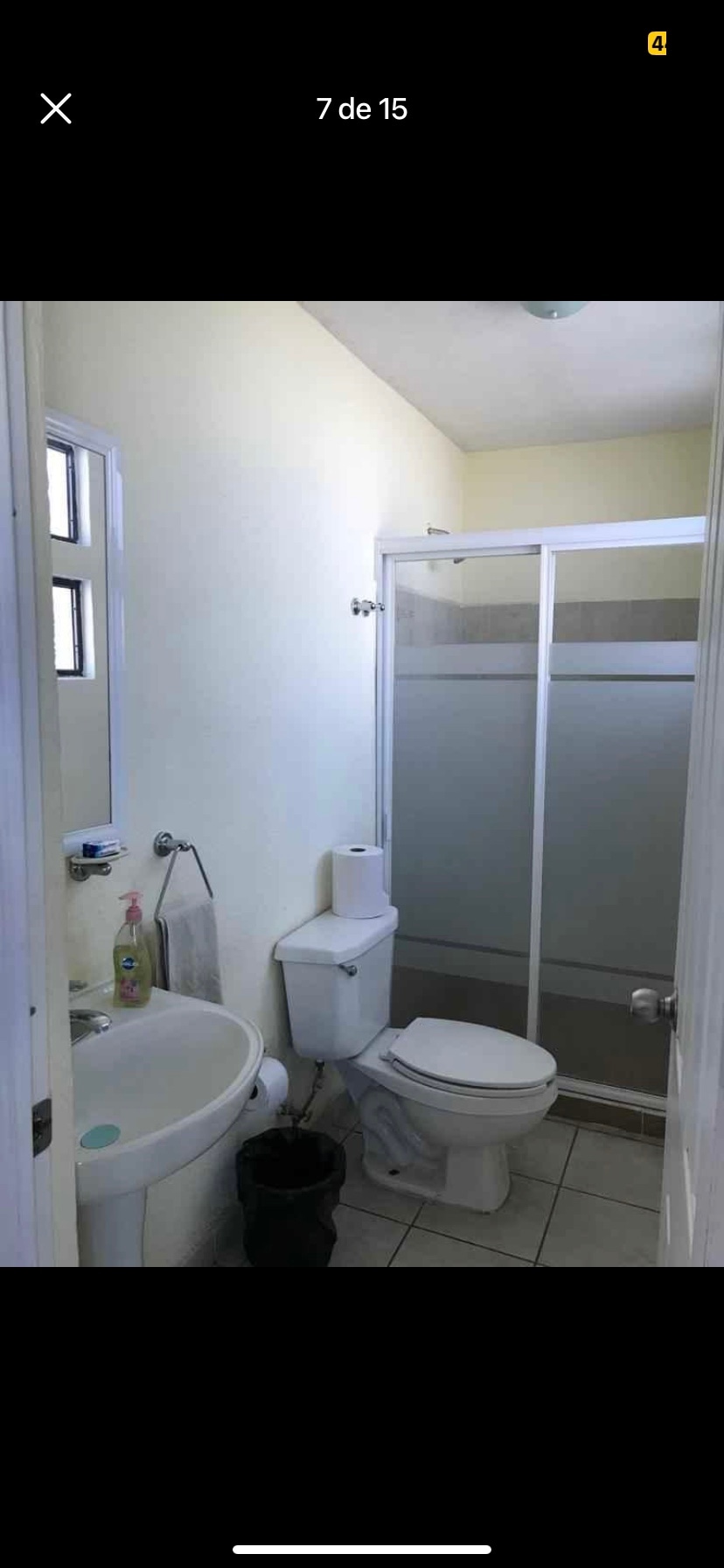A clean and functional bathroom features white walls and tiled flooring. A shower enclosure with sliding glass doors is visible alongside a compact sink and toilet. Natural light is provided by two small windows, enhancing the space's brightness.