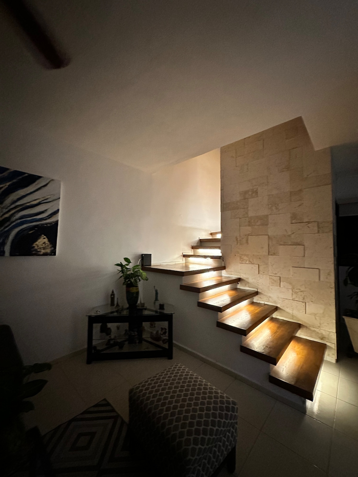 A staircase is illuminated softly by lights within the wooden steps, creating a gentle ambiance. A stone wall adds texture to the space, while a piece of abstract art adorns the wall nearby. A small table with decorative items is positioned at the base of the stairs.