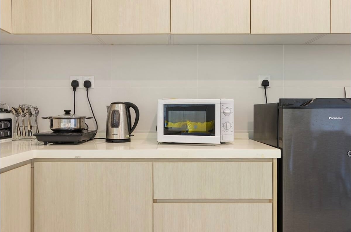 The kitchen area features a countertop with a microwave, electric kettle, and a small pot on the stove. A refrigerator is positioned on the right, while cabinetry provides additional storage. Sleek, minimalist cabinetry in light wood creates a clean and functional space.