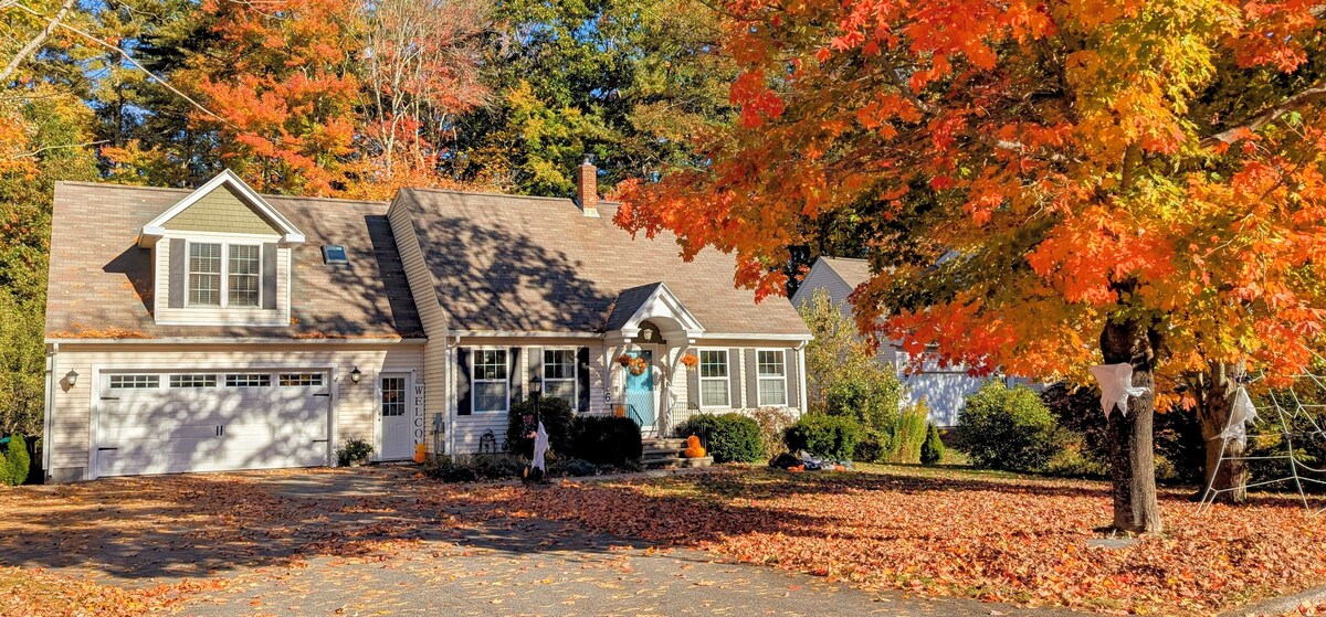 A charming home is surrounded by vibrant autumn foliage and a carpet of fallen leaves. The inviting exterior features a garage and large windows, while a colorful tree adds warmth to the scene. A playful atmosphere is enhanced by nearby decorations.