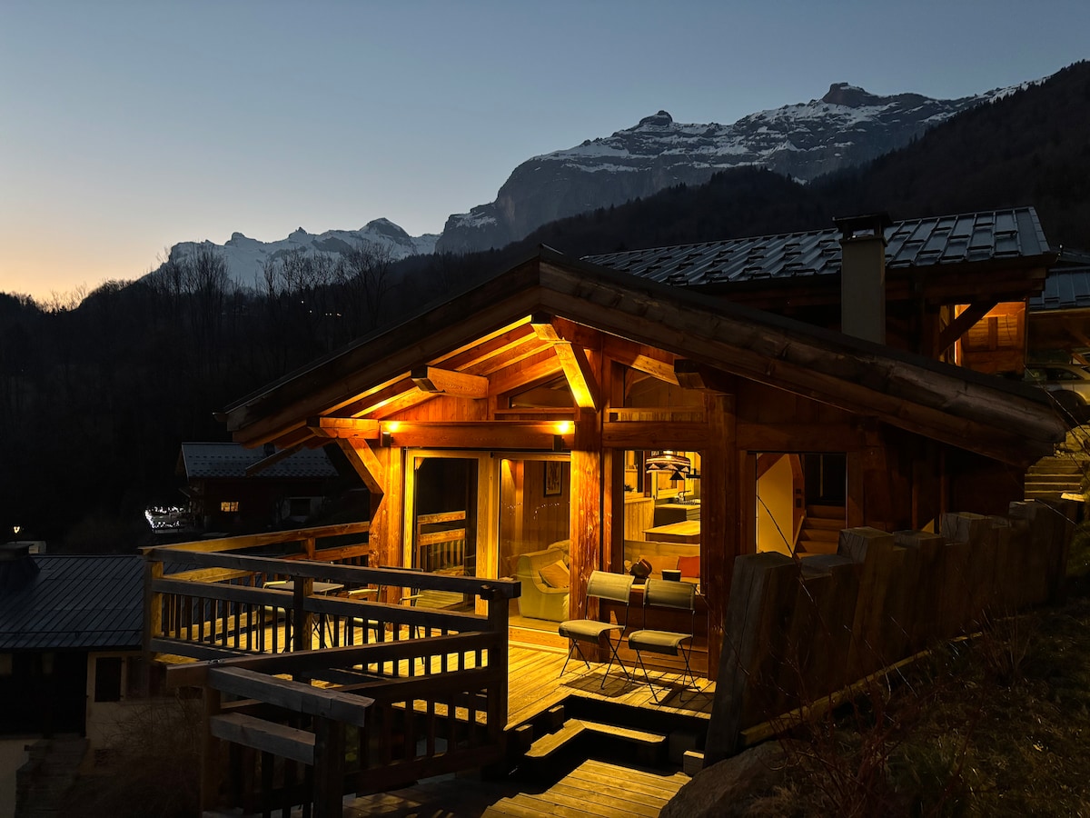 The chalet is illuminated in soft amber tones as dusk settles, showcasing its wooden façade and spacious balcony. Snow-capped mountains are visible in the background, with a cozy seating area positioned on the deck, creating an inviting outdoor space.