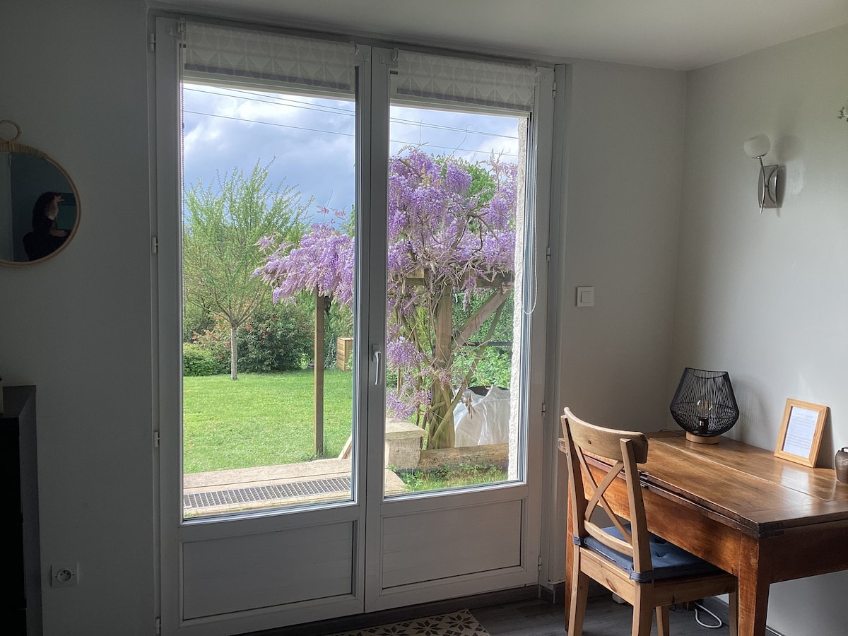 A window view showcases a vibrant garden with a blooming purple tree. A wooden table and chair are situated near the glass doors, inviting natural light into the space. The surrounding greenery provides a tranquil retreat, enhancing the calm atmosphere of the room.