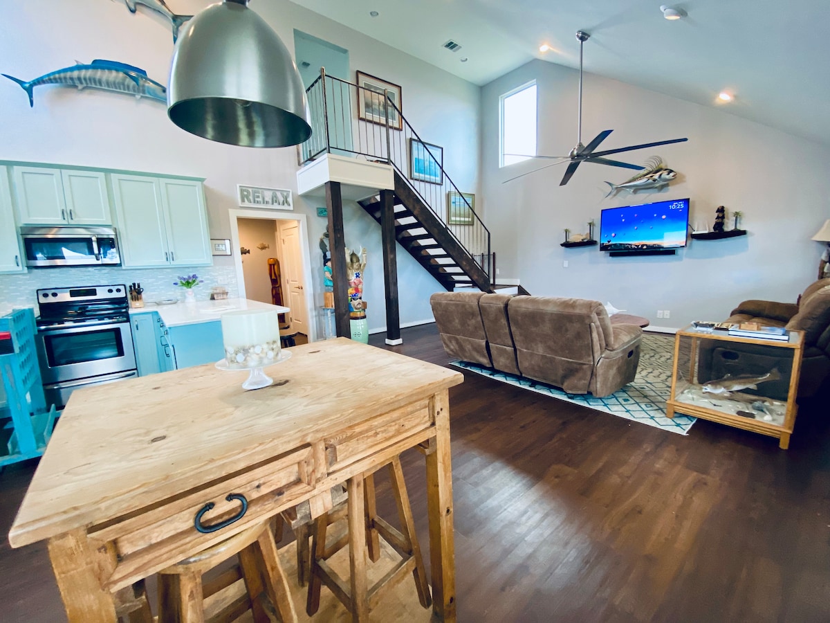 An open-concept living area features a spacious kitchen equipped with stainless steel appliances and a rustic wooden island. A comfortable seating arrangement, including a large couch and a coffee table, is visible. Natural light floods the space through a large window, complementing the inviting decor.