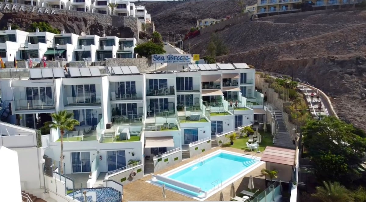 Aerial view of a modern apartment complex features a swimming pool surrounded by a terrace. The building showcases multiple units with balconies facing the pool and valley. The area is bordered by rugged terrain and other nearby structures.