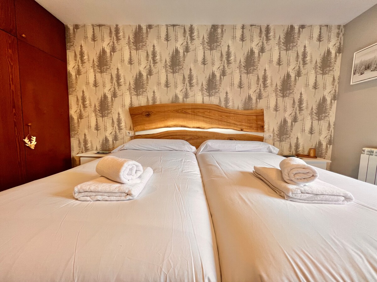 The image showcases a sleeping area featuring two spacious beds, arranged side by side with neutral bedding. Freshly rolled towels are placed atop each bed. A wooden headboard adds a natural touch, and a subtle forest-themed wallpaper lends a calming backdrop.