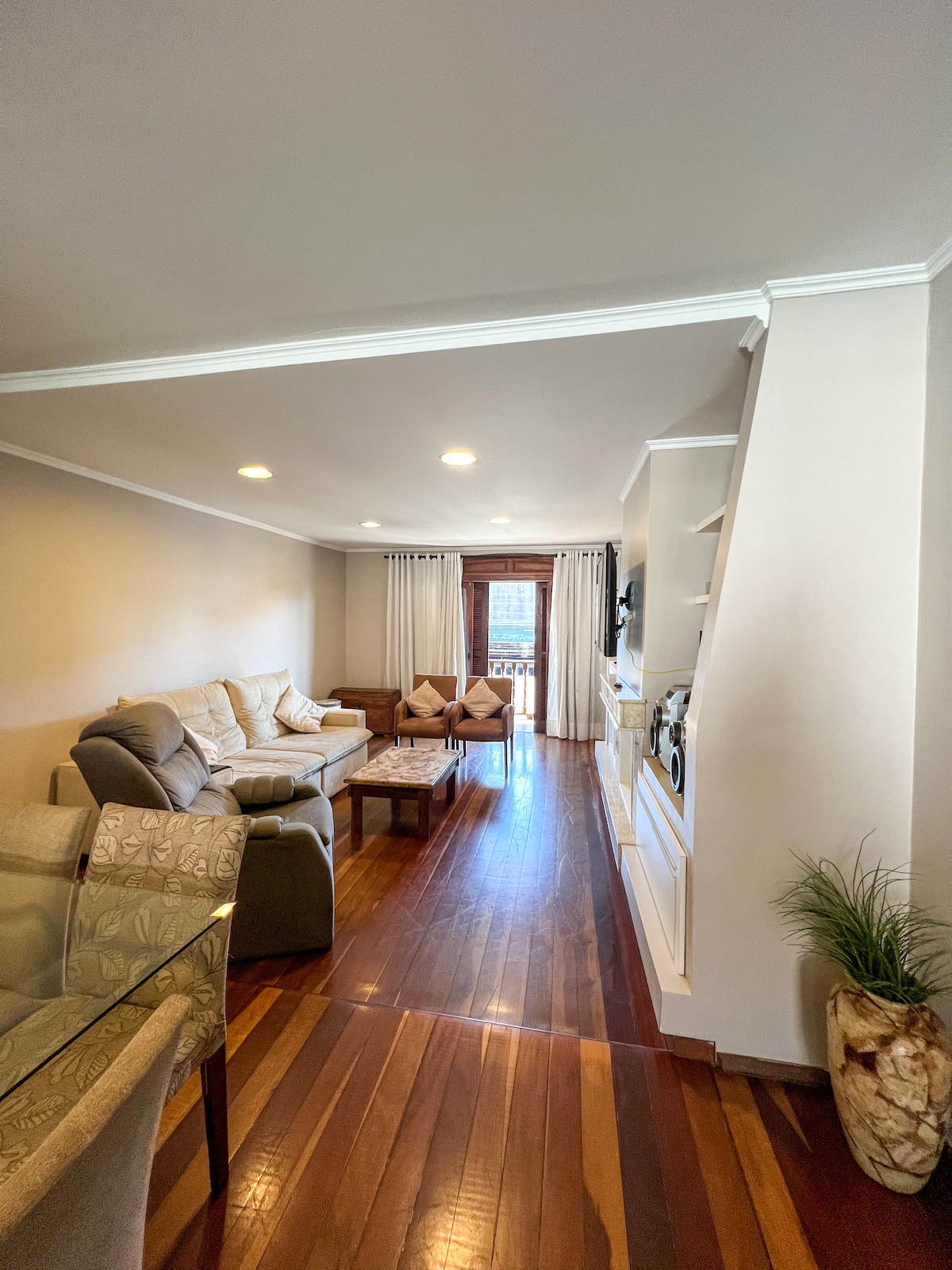 The living area features a comfortable seating arrangement with a light-colored sofa and pillows, accompanied by a wooden coffee table. A dining area, with a transparent table, is positioned nearby. Sunlight enters through the glass door, which leads to a small balcony.