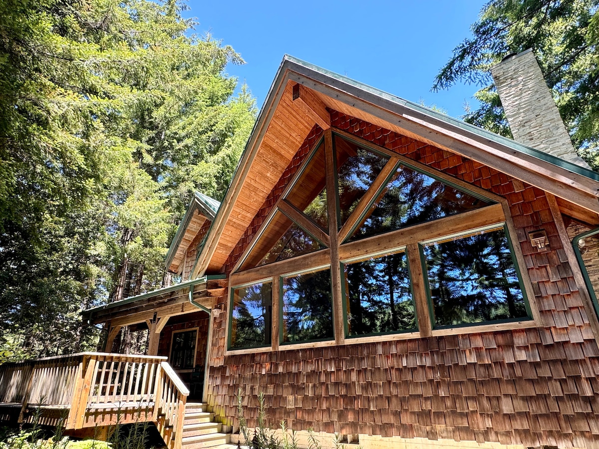 The exterior of the lodge is highlighted by its unique architecture, featuring a combination of wooden shingles and large picture windows. The sloped roof adds character, and a staircase leads to the entrance, surrounded by verdant trees that contribute to a secluded atmosphere.