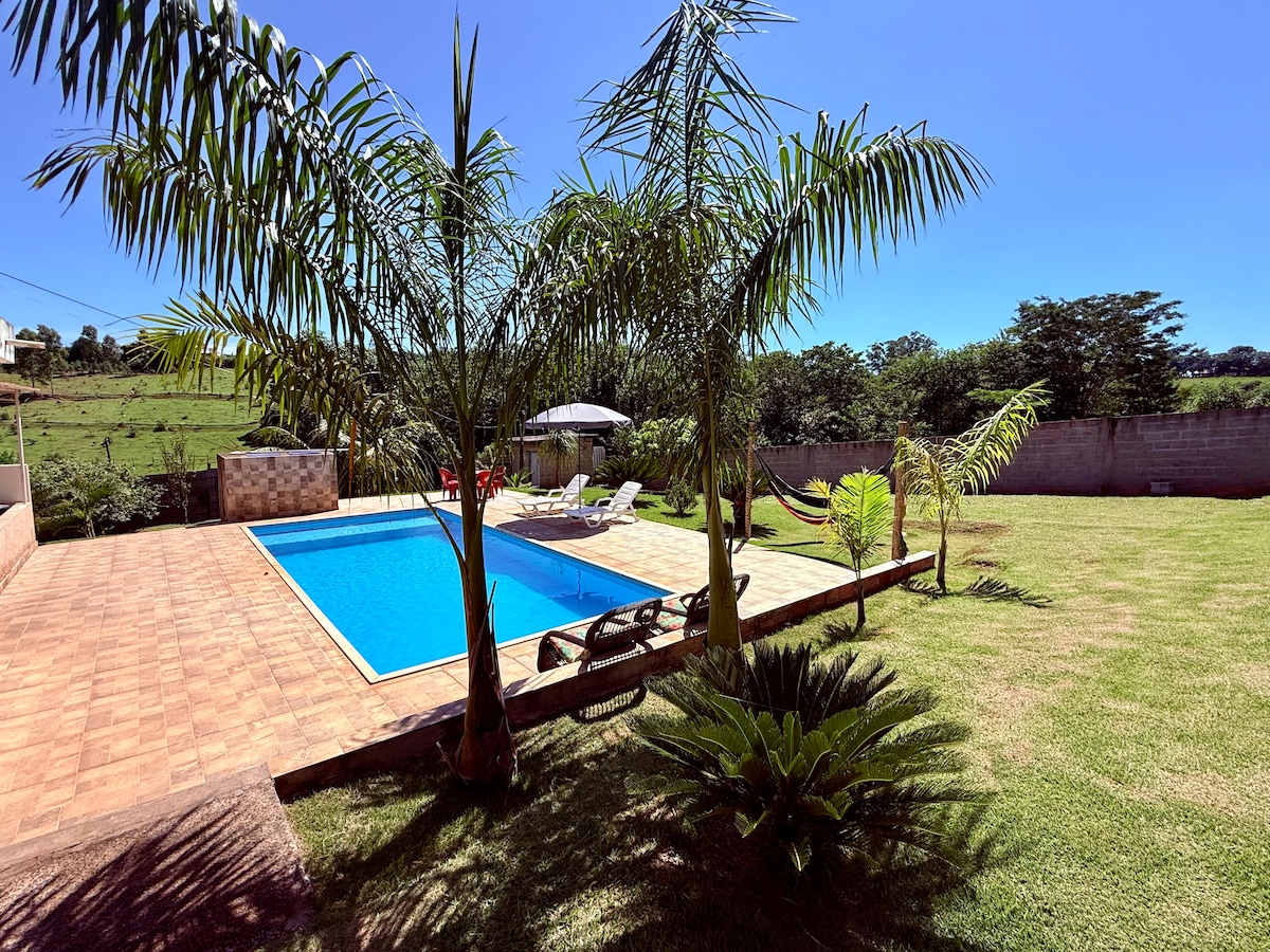 The image showcases a bright outdoor space featuring a refreshing swimming pool surrounded by lush greenery. Sun loungers are positioned near the pool, and an umbrella provides shaded seating. The well-maintained lawn offers a spacious area for relaxation and outdoor activities.
