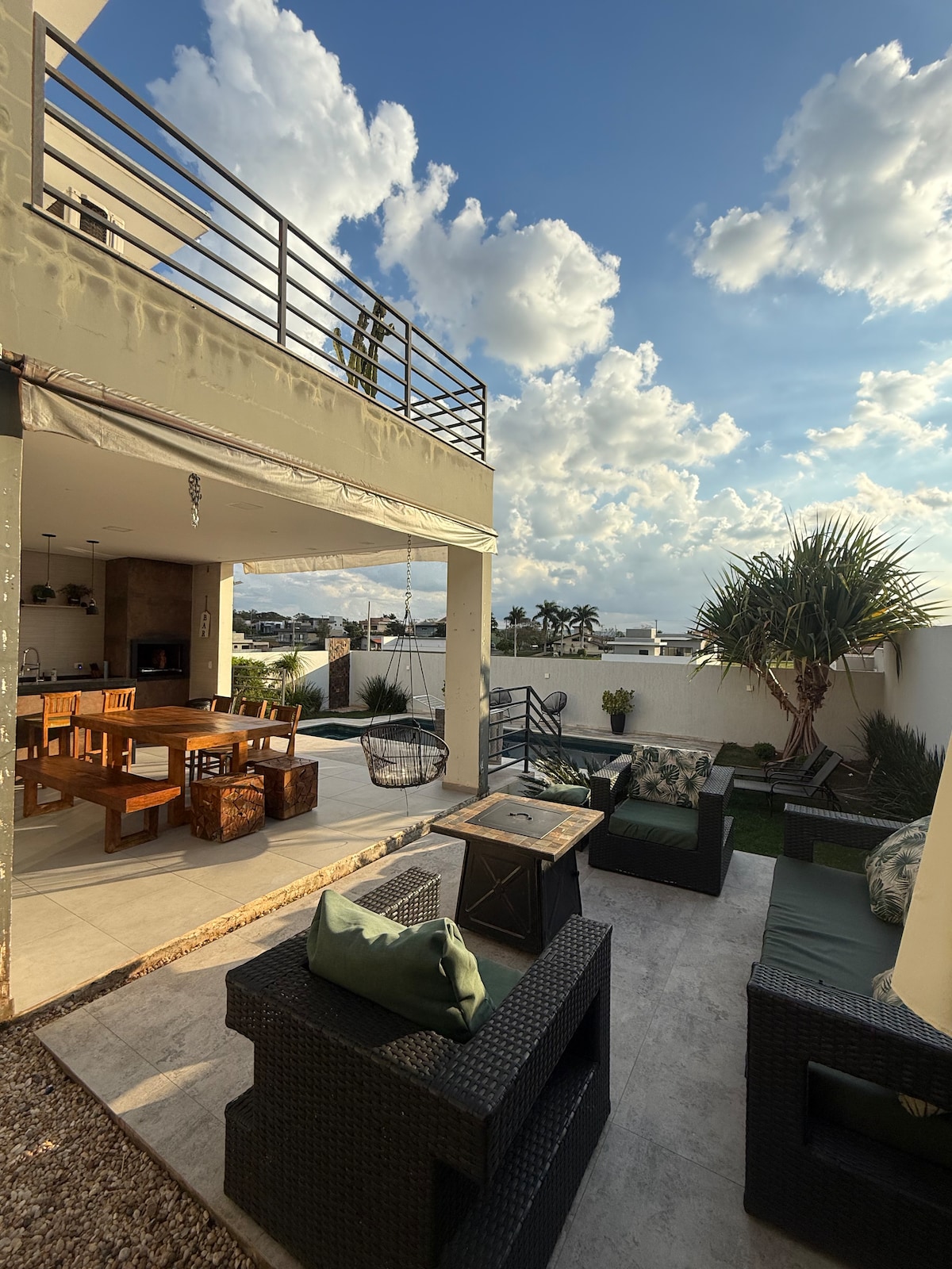 An outdoor seating area is featured, with comfortable rattan chairs and a fire pit, surrounded by lush greenery. A spacious dining table is visible nearby, under a blue sky dotted with fluffy clouds. The area invites relaxation and outdoor dining.