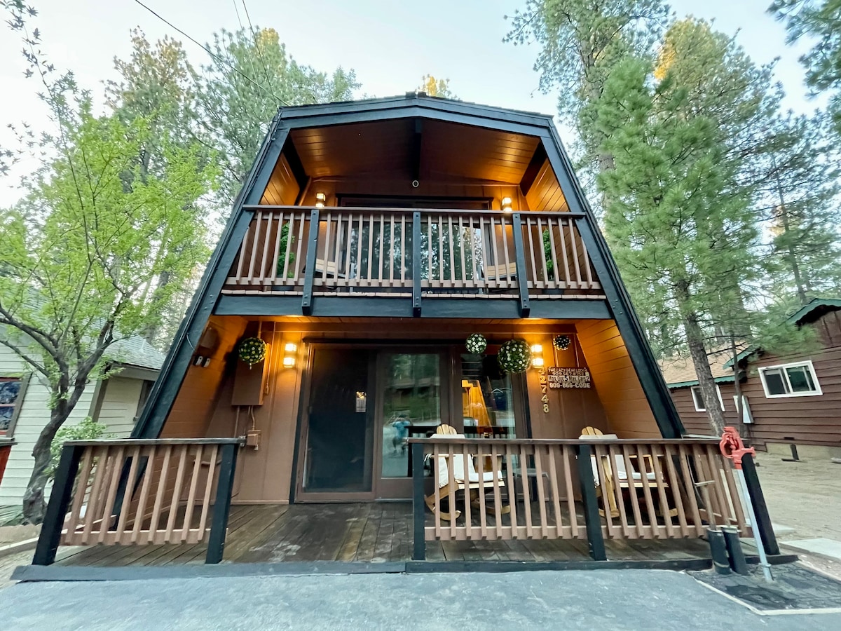 The charming cabin features a unique A-frame structure with a dark exterior and wooden accents. A spacious front porch, adorned with two hanging flower arrangements, invites relaxation, while the upper balcony offers seating and a view of the surrounding pine trees.