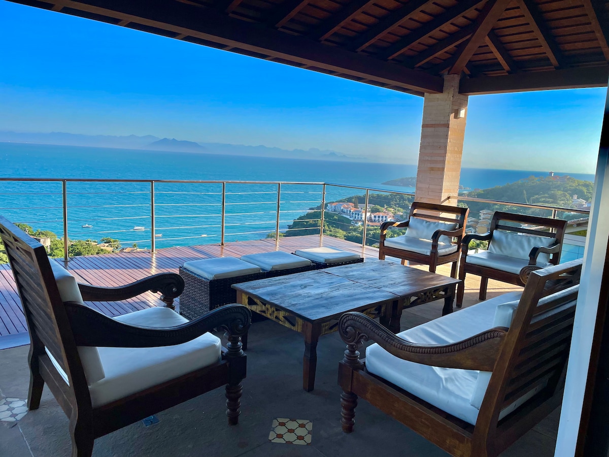 A spacious balcony area features a wooden table surrounded by four comfortable chairs with white cushions. The backdrop showcases panoramic ocean views under a clear blue sky, providing a serene and inviting outdoor space.