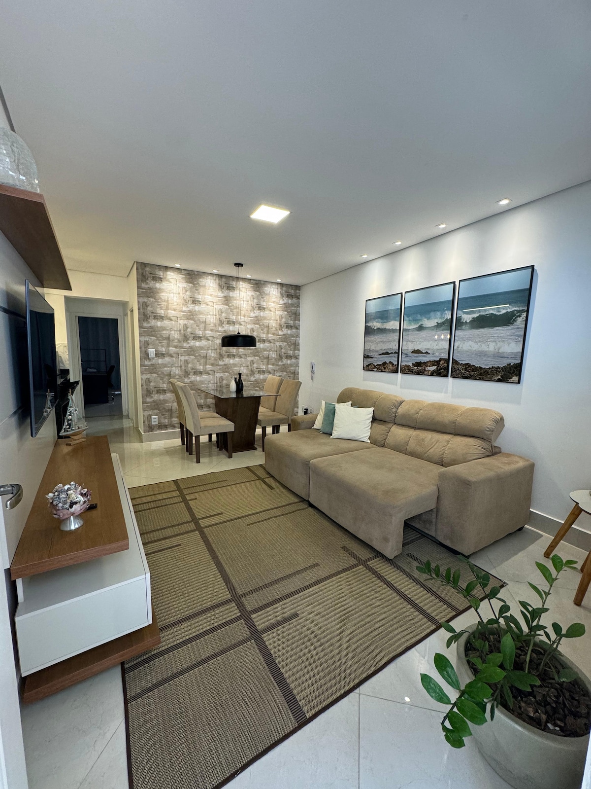 A modern living area features a comfortable beige sofa facing a sleek entertainment unit. A round dining table with two chairs is placed nearby. The space is enhanced by three framed ocean prints on the wall and a green plant positioned on the floor.