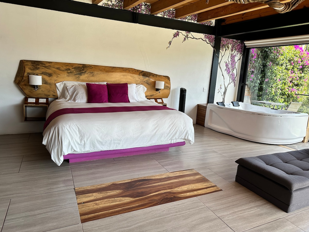 A spacious bedroom features a king-size bed with a unique wooden headboard and vibrant purple accents. Natural light pours in through large glass doors, leading to a private terrace with views of lush greenery. A luxurious jacuzzi adds to the inviting atmosphere.