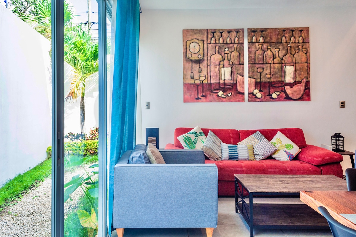 A living area features a red sectional sofa adorned with colorful cushions, accompanied by a gray armchair. Two framed artworks hang above the sofa. Large glass doors provide natural light and lead to a garden area, enhancing the indoor-outdoor connection.
