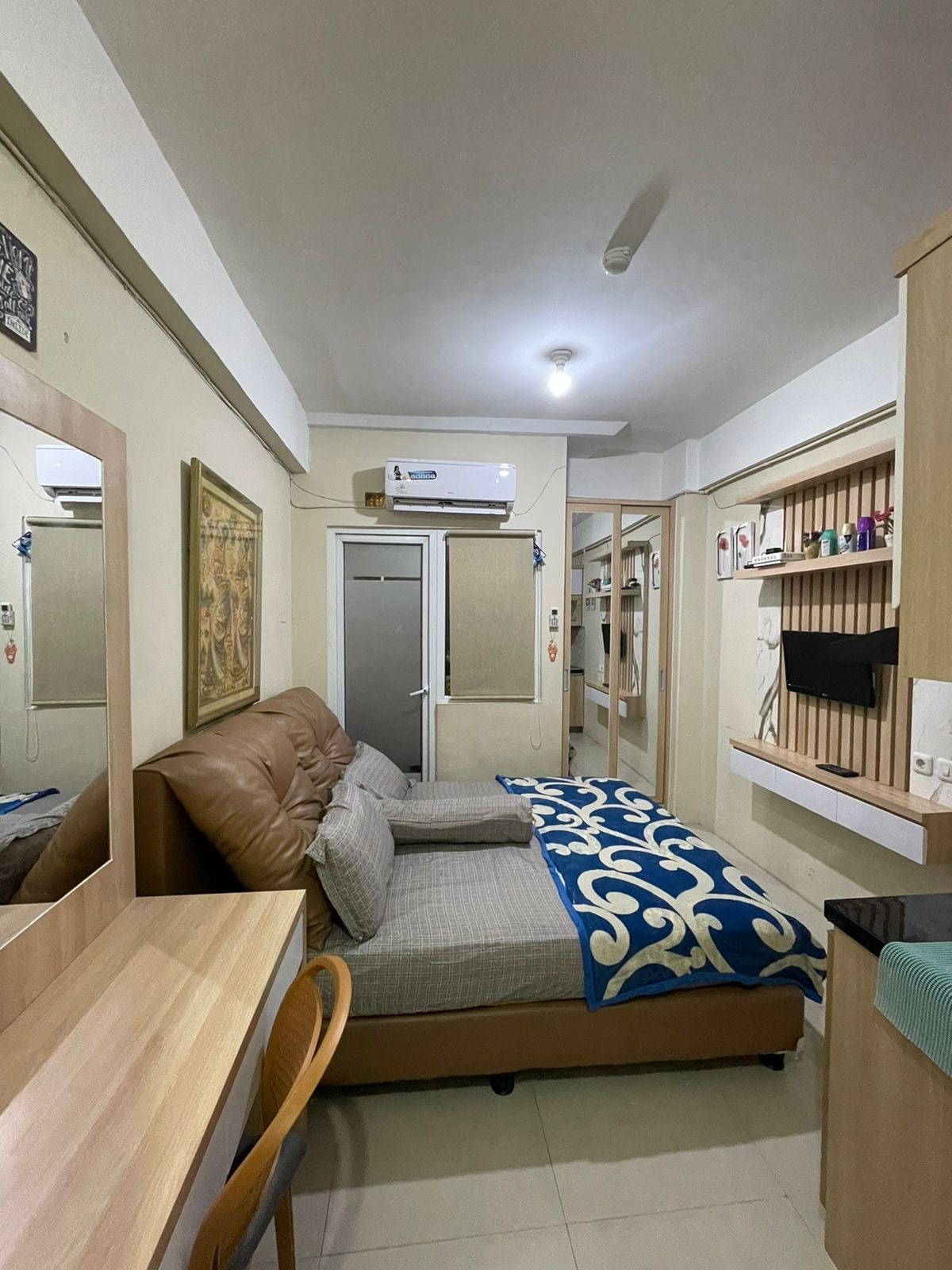 The studio room features a spacious bed with a decorative blanket. A mirror and a work desk are positioned against the wall, while a wall-mounted TV is visible. An air conditioning unit is situated above, and sliding doors lead to an enclosed space.