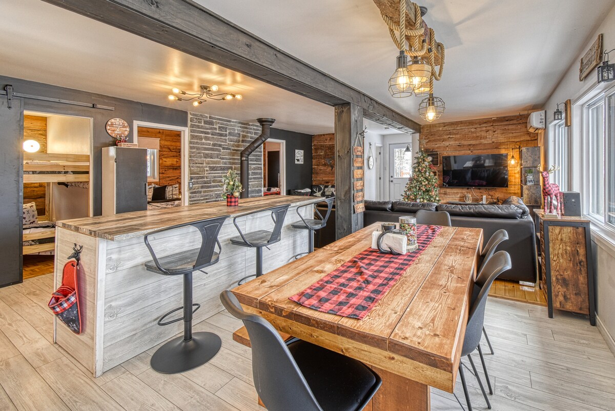 An open-concept living space showcases a rustic wood dining table surrounded by sleek black chairs. A kitchen island with bar stools is visible, along with modern appliances and a cozy seating area with a television. Natural light fills the room, highlighted by wooden accents.