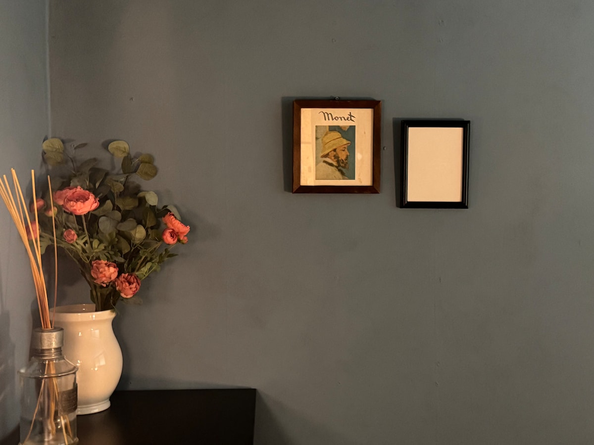 A corner of the room features a wooden-framed artwork titled 'Mount' alongside an empty black frame. A white vase holds pink flowers, and a glass jar with reeds adds a decorative touch to the dark surface below.