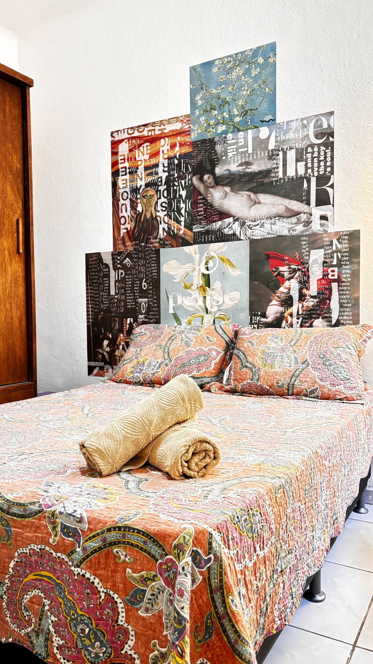 A cozy sleeping area features a double bed with a patterned orange and beige bedspread. Decorated walls display a collage of vibrant artworks. Soft towels are neatly arranged at the foot of the bed, contributing to the clean and organized atmosphere.