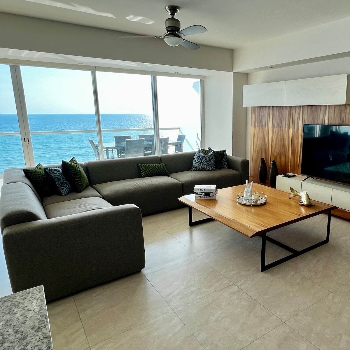 A contemporary living area features a spacious sectional sofa adorned with decorative cushions. A coffee table sits in front, with a Smart TV mounted on the wall, overlooking large glass doors that open to a balcony with a view of the ocean.