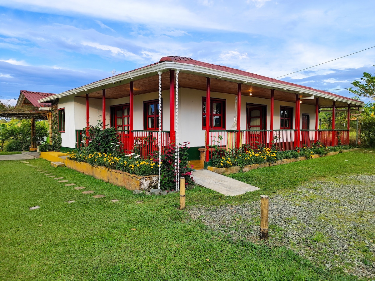 The exterior of Casa Laureles showcases a charming single-story structure with a red and white color scheme. A covered porch extends across the front, framed by vibrant flowers and greenery. The surrounding lawn offers a spacious area for relaxation and enjoyment.