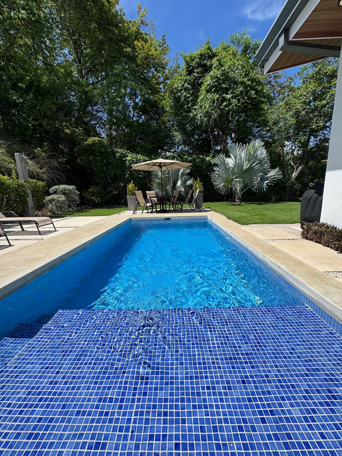 A private swimming pool is surrounded by lush greenery and a well-maintained garden. Patio furniture with an umbrella is positioned at one end of the pool, creating a relaxing space for outdoor gatherings. Sunlight reflects beautifully on the water's surface.