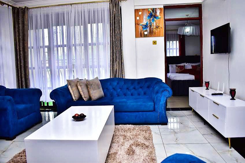 A bright living area features a light-colored rug beneath a white coffee table, surrounded by a plush blue sofa and matching armchairs. A wall-mounted TV is positioned opposite the seating, while large windows allow natural light to fill the space.