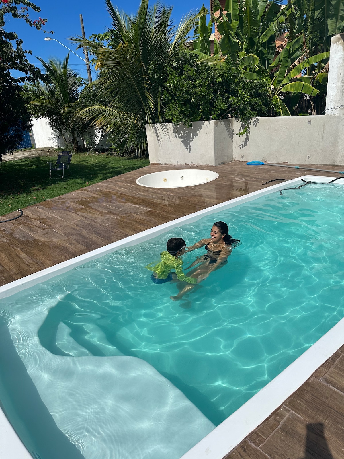 A swimming pool with clear turquoise water is surrounded by lush green palm trees and manicured grass. A spacious deck area features a sunken circular seating area. Natural sunlight illuminates the scene, creating a relaxed outdoor ambiance.