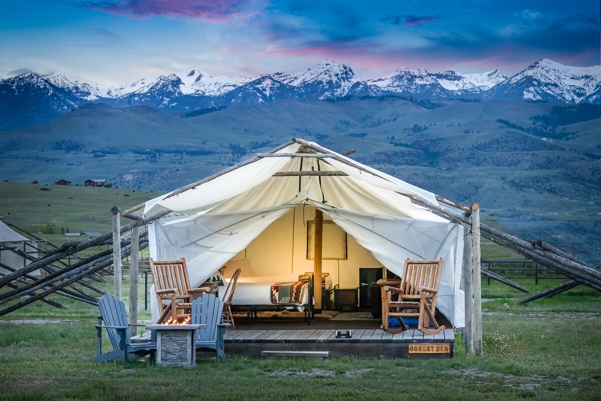 A tent with an open front displays comfortable seating on a private deck. Adirondack chairs are positioned around a small fire table, offering a cozy space to enjoy the surrounding landscape. Majestic mountains are visible in the background, illuminated by soft evening light.