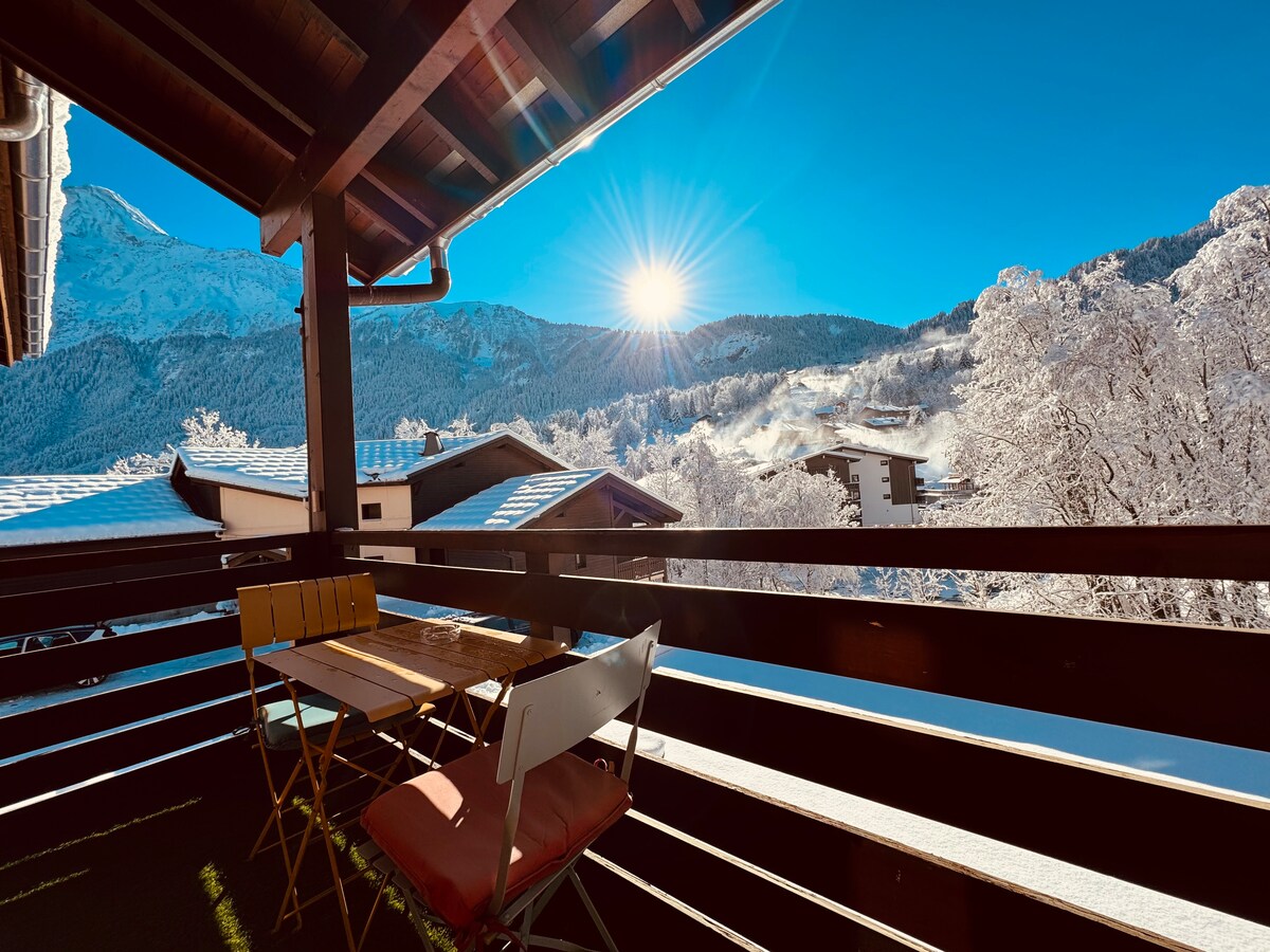 A balcony is showcased with a small table and chairs, surrounded by snow-covered trees and mountain views. The bright sun illuminates the scene, enhancing the serene winter atmosphere.