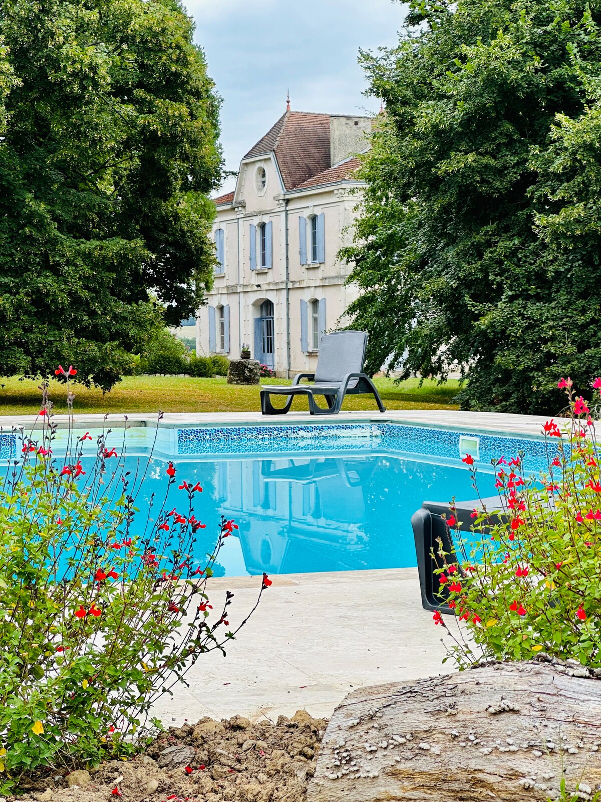 A clear blue swimming pool is framed by colorful flowers and lush greenery. A sun lounger is positioned beside the pool, providing a spot for relaxation. The exterior of the spacious house is visible in the background, featuring white walls and blue shutters.