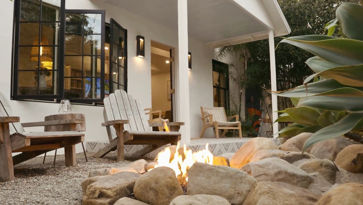 An inviting outdoor seating area is highlighted by comfortable wooden chairs arranged around a fire pit. Flickering flames illuminate smooth stones, with the serene ambiance enhanced by lush greenery and the cozy home facade visible in the background.