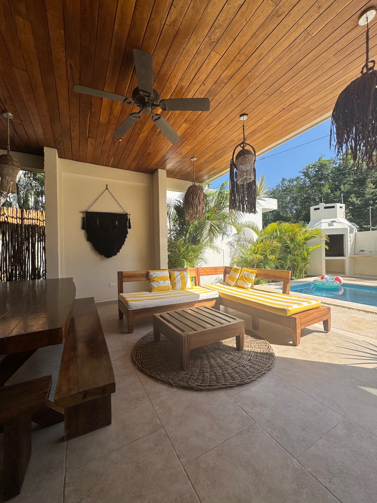 A covered outdoor seating area features two lounge chairs draped in yellow and white stripes, accompanied by a central coffee table. A wooden dining table is visible, with a ceiling fan and light fixtures made from natural materials above. Lush green plants and a pool are seen in the background.